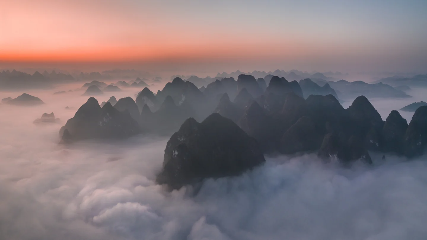 Low clouds wrap around dark karst peaks beneath a pale pink horizon.