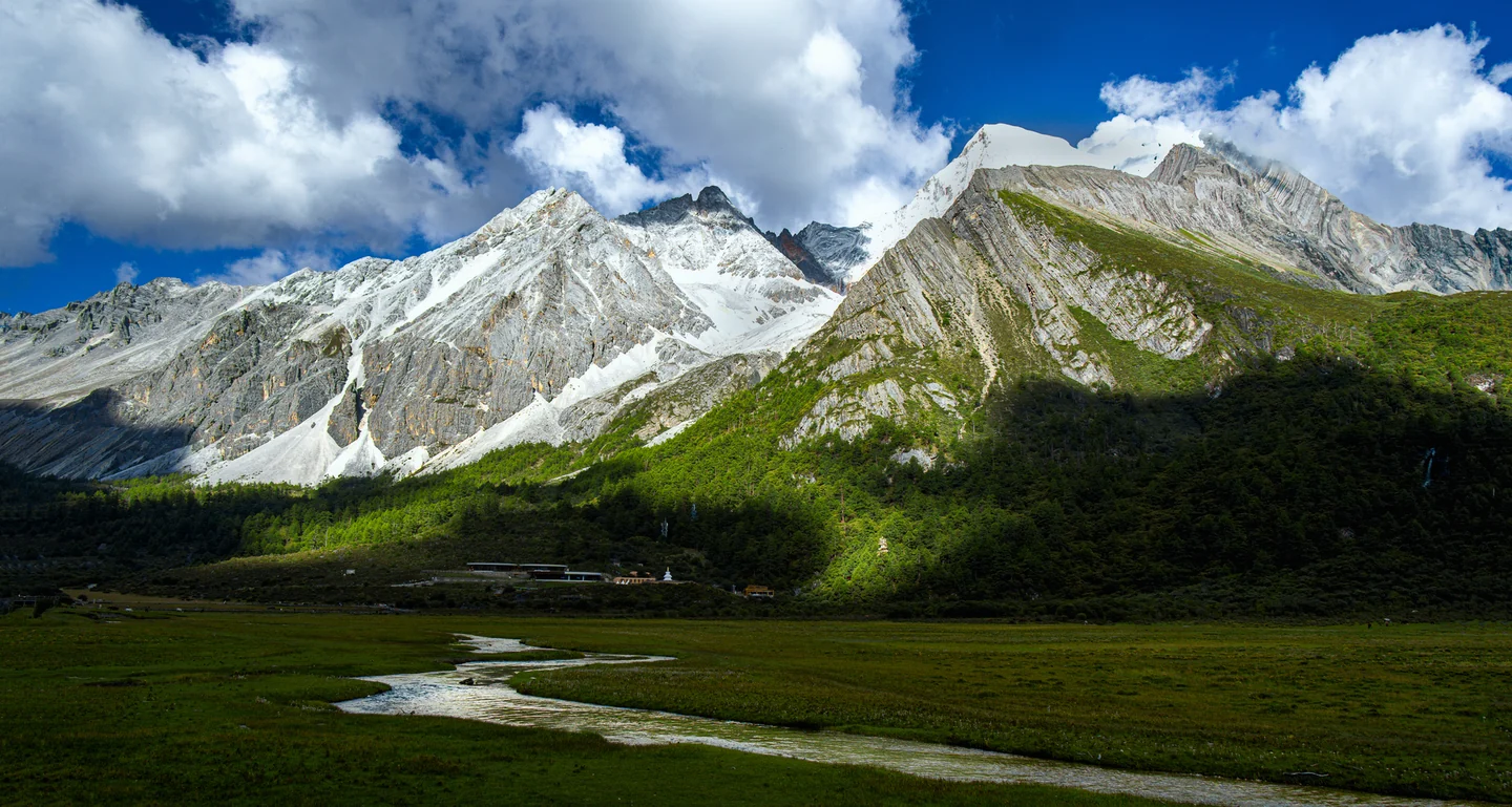 A stream winds across a broad green pasture beneath snow-covered peaks.