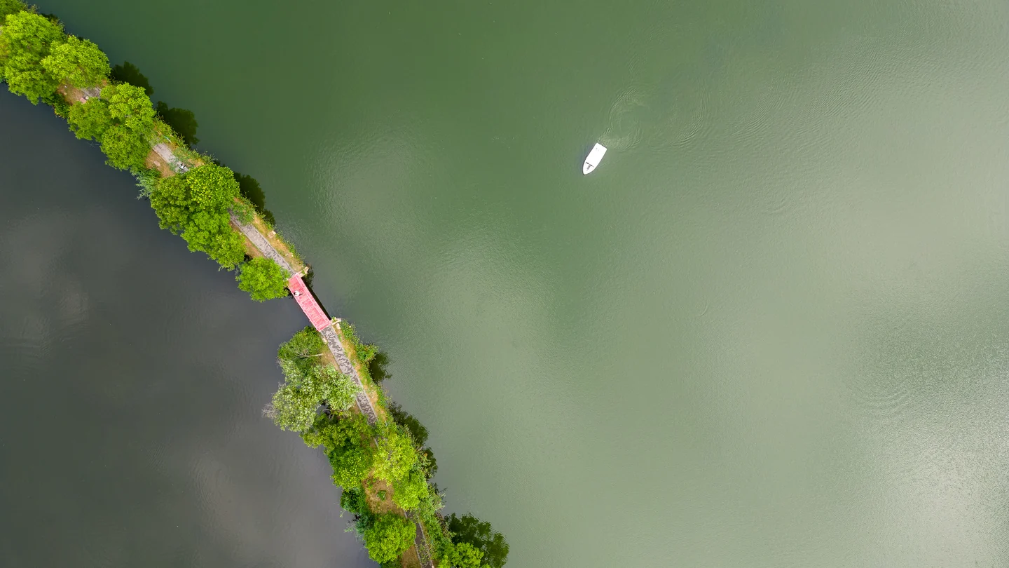 A small white boat floats beside a tree-lined path across green water, seen from above.