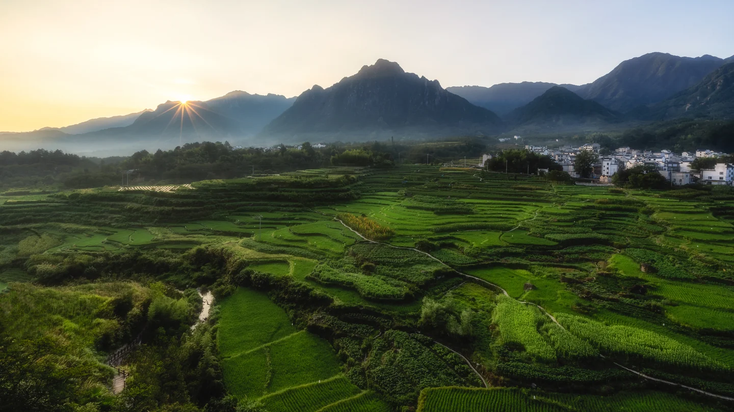Sunlight breaks over distant mountains and a patchwork of green terraced fields.